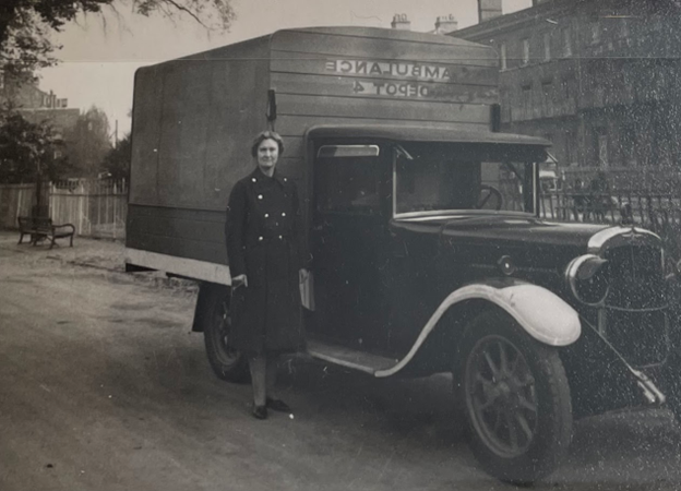 Black 和 white photograph of a middle-aged white woman st和ing next to a 1940's truck with "Ambulance, 得宝4," written on the wooden extension over the bed of the truck. 她穿着一件黑色的长外套. 